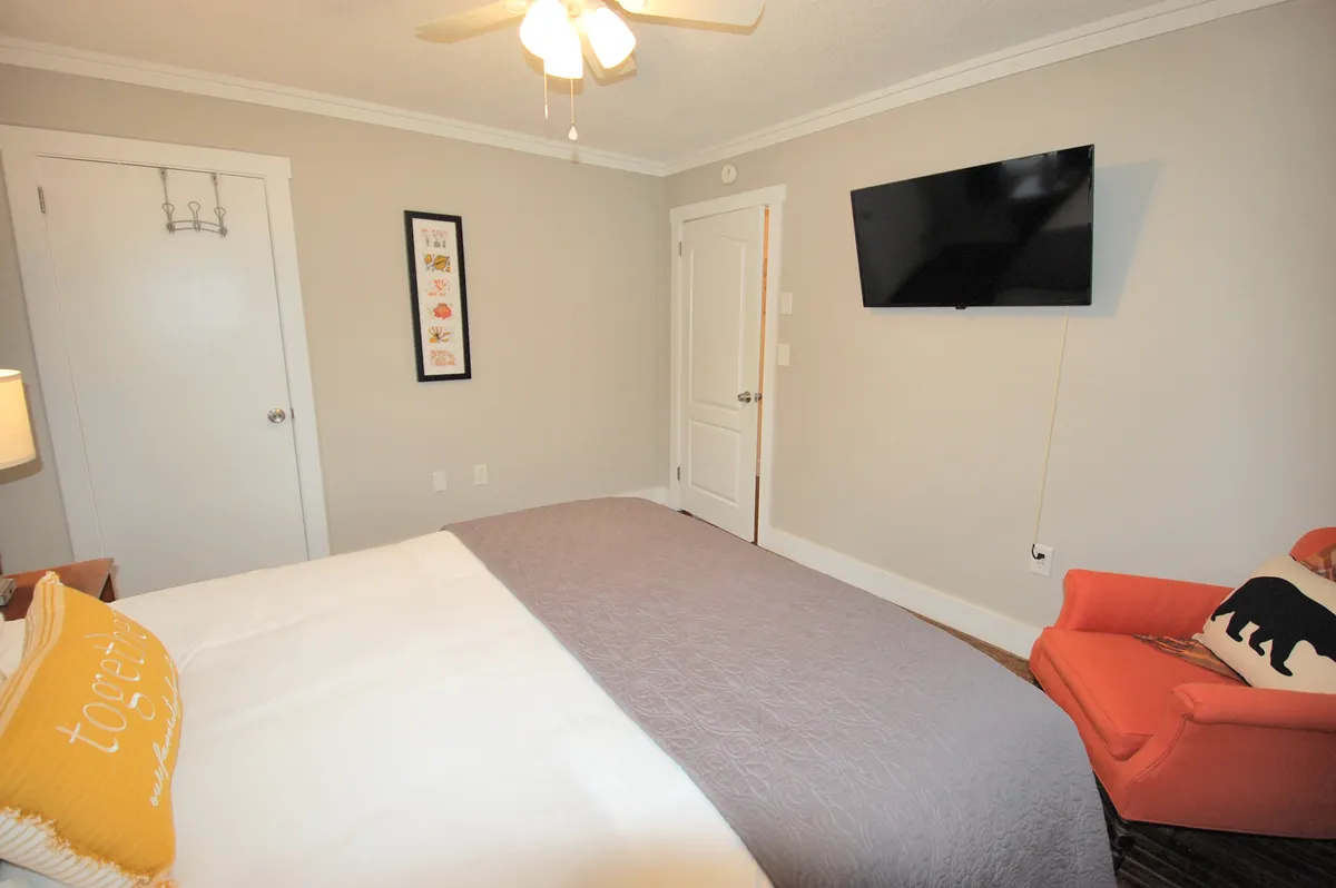 A comfortable bedroom scene showcasing a bed with decorative pillows, an orange armchair perfect for reading or relaxing, a wall-mounted TV, and framed artwork that adds a homely touch. The lighting feels soft, enhancing the room's warmth and comfort.