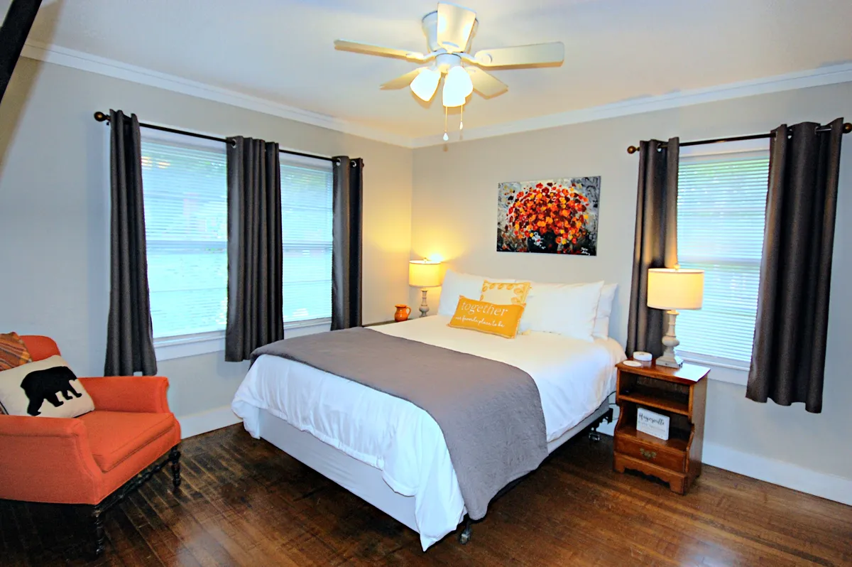 A welcoming bedroom scene featuring a comfortable bed with fresh linens, an armchair beside a large window, a stylish lamp, and thoughtfully curated wall art. The space receives soft natural light, creating a cozy spot for reading or relaxing after a day out.
