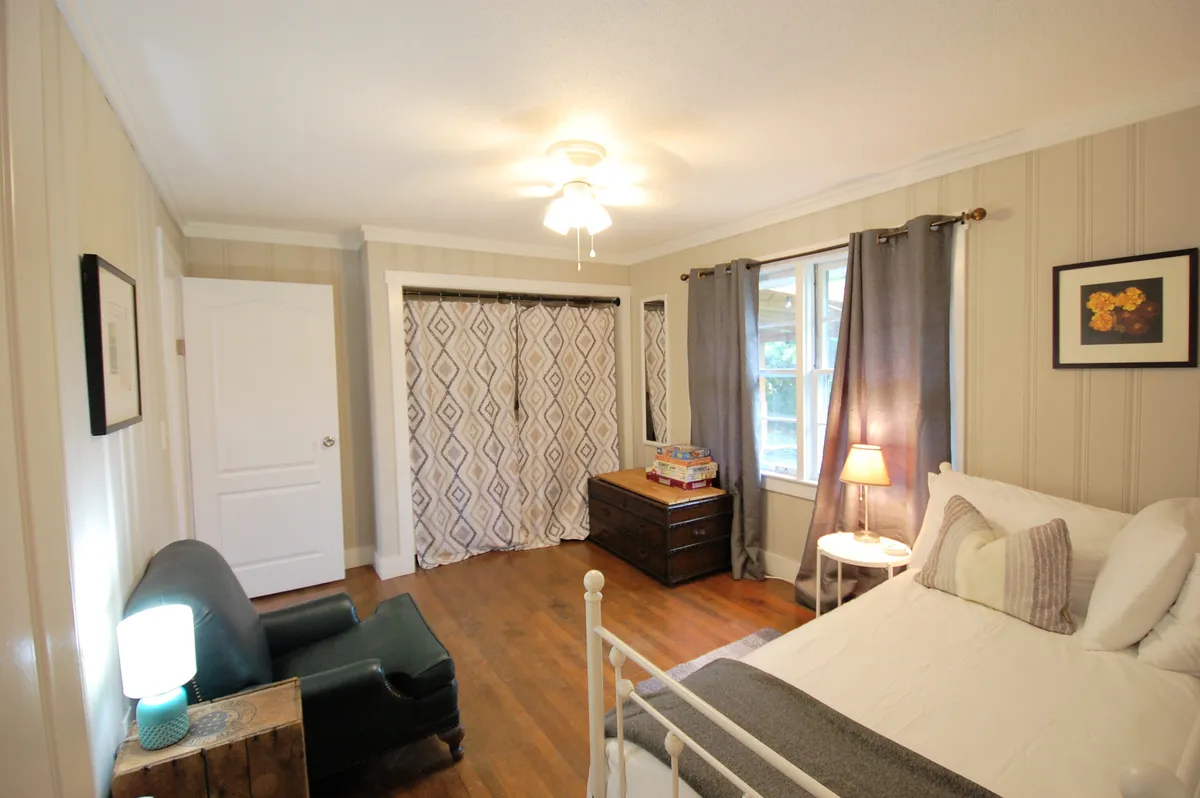 A welcoming bedroom filled with natural light, featuring a comfortable bed with crisp white bedding, a green armchair near a window, and decorative curtains framing a view outside. The nightstand and lamp add a homey touch, and the window hints at the presence of fresh air and a connection to the outdoors.