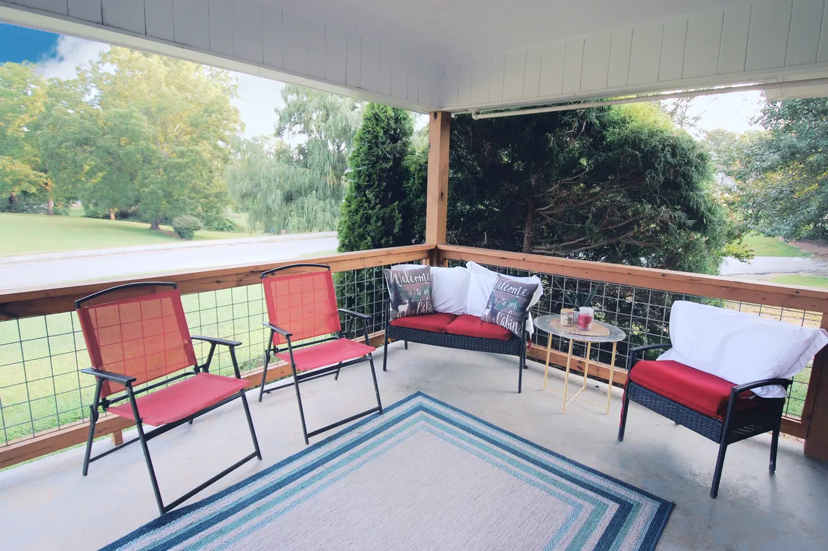 A charming exterior shot of the front porch featuring two vibrant red chairs with welcome pillows, a small table, and a cozy area rug. The porch is bathed in natural light, evoking a sense of invitation and relaxation in a quiet neighborhood setting.