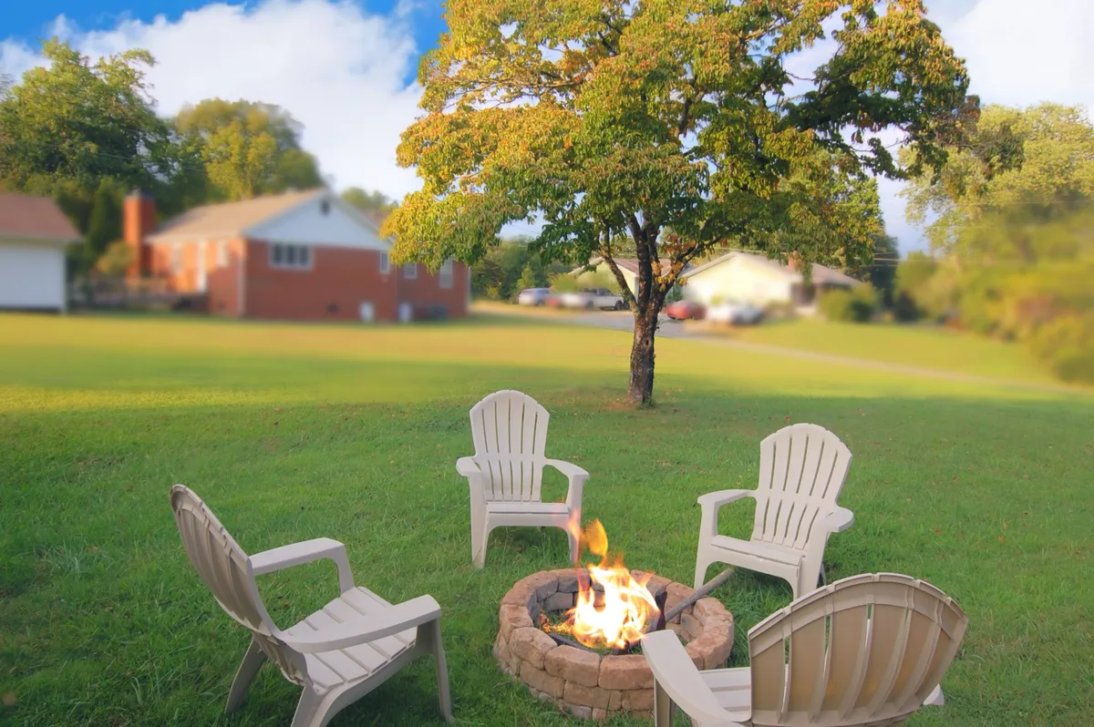 An inviting backyard fire pit area with Adirondack chairs arranged around the fire, surrounded by trees and neighboring houses in the background. The setting is perfect for unwinding and sharing stories after a day of adventure.