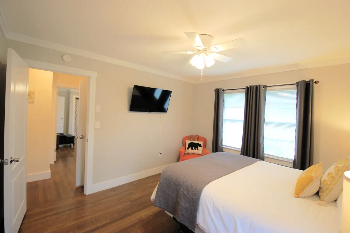 This image features a cozy bedroom with a bed adorned with decorative pillows, a wall-mounted flat-screen TV, a chair with a playful bear motif, and a window letting in soft natural light. The nightstand is visible next to the bed, offering a convenient spot for essentials. The overall ambiance is tranquil and inviting.